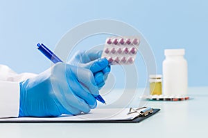 Doctor`s hands in blue medical gloves write a prescription and hold a blister of pills on a blue background. concept of medicine,