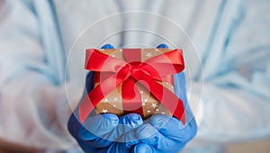 Doctor`s hands in medical gloves holds a gift box with a red ribbon. Clse-up