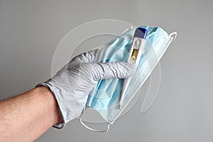 The doctor`s hands protected by blue gloves hold a blue surgical mask and fever thermometer on gray background