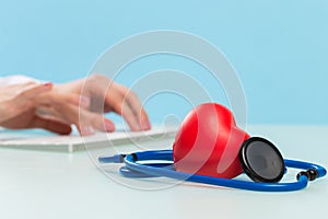 Doctor`s hands are typing on a white keyboard, next to them are a dark blue stethoscope and a red heart on a blue background.