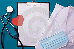 A doctor`s uniform lies on the table, a notebook for notes, a red heart, a mask and a stethoscope lie nearby