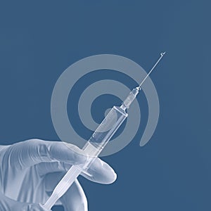 Doctor`s white-gloved hand holding a syringe with transparent liquid isolated on blue background.
