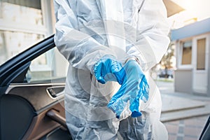 Doctor, scientist or nurse wearing face mask and biological hazmat protective suit putting on rubber gloves near car outdoor.