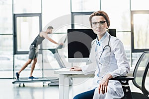 doctor sitting at desk near sportsman running on treadmill during endurance test in gym.