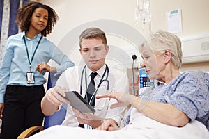 Doctor Sitting By Female Patient's Bed Using Digital Tablet