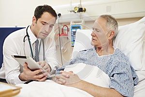 Doctor Sitting By Male Patient's Bed Using Digital Tablet