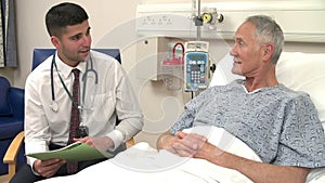 Doctor Sitting By Senior Male Patient's Bed In Hospital