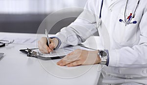 Doctor sitting and working with clipboard of medication history record in clinic at his working place, close-up. Young