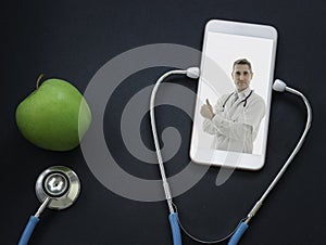 Doctor on smartphone screen, stethoscope and green apple on table, doctor online concept, online medical communication and medical