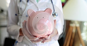 The doctor standing in the clinic holds a pink piggy bank