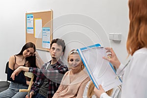 A doctor standing in front of her office reads out the next person and invites them in for a check-up. Close-up shot