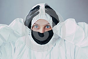 Doctor standing on a gray background dressed in a viral protective suit and black mask holding hands in black medical gloves behin