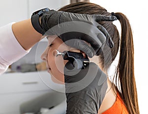 A doctor in sterile black medical gloves pierces the ears of a young girl in the medical office with a piercing gun.