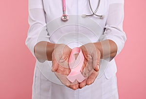 Doctor with stethoscope holding pink ribbon on color background, closeup. Breast cancer awareness