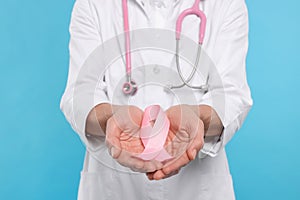 Doctor with stethoscope holding pink ribbon on light blue background, closeup. Breast cancer awareness