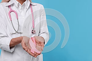 Doctor with stethoscope holding pink ribbon on light blue background, closeup and space for text. Breast cancer awareness