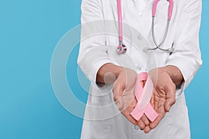 Doctor with stethoscope holding pink ribbon on light blue background, closeup and space for text. Breast cancer awareness