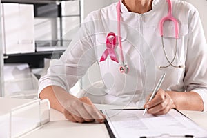 Doctor with stethoscope and pink ribbon at white desk indoors, closeup. Breast cancer awareness