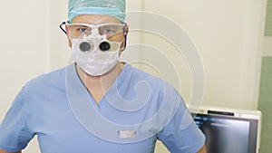 Doctor takes off magnifying binocular glasses and sterile mask, looks into camera and smiles
