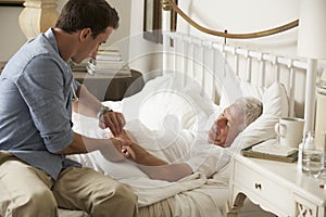 Doctor Taking Pulse Of Senior Male Patient In Bed At Home