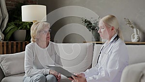 A doctor talking to an elderly lady patient sitting on a sofa in an apartment