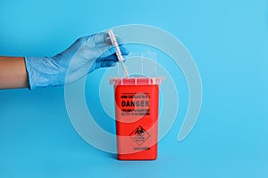 Doctor throwing used syringe into sharps container on light blue background, closeup