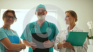 Doctor and two nurses are posing in front of the camera. Doctors laugh standing in front of the window and holding