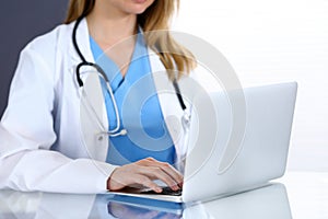 Doctor typing on laptop computer while sitting at the glass desk in hospital office. Physician at work. Medicine and
