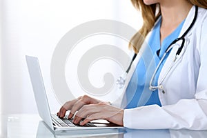 Doctor typing on laptop computer while sitting at the glass desk in hospital office. Physician at work. Medicine and