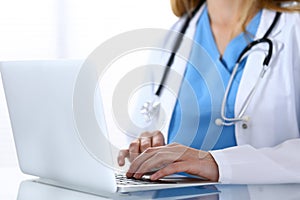 Doctor typing on laptop computer while sitting at the glass desk in hospital office. Physician at work. Medicine and