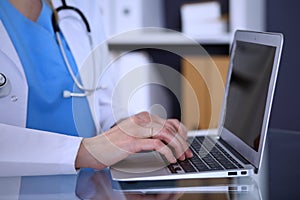 Doctor typing on laptop computer while sitting at the glass desk in hospital office. Physician at work. Medicine and