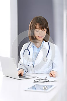 Doctor typing on laptop computer while sitting at the white table in hospital office. Physician at work. Medicine and