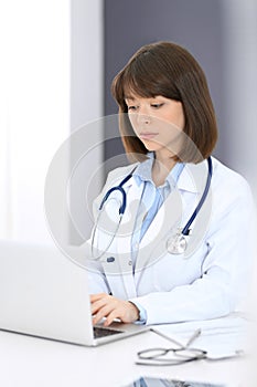 Doctor typing on laptop computer while sitting at the white table in hospital office. Physician at work. Medicine and