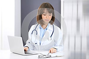 Doctor typing on laptop computer while sitting at the white table in hospital office. Physician at work. Medicine and