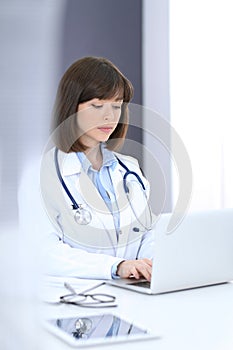 Doctor typing on laptop computer while sitting at the white table in hospital office. Physician at work. Medicine and