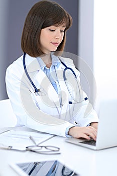 Doctor typing on laptop computer while sitting at the white table in hospital office. Physician at work. Medicine and