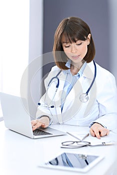 Doctor typing on laptop computer while sitting at the white table in hospital office. Physician at work. Medicine and