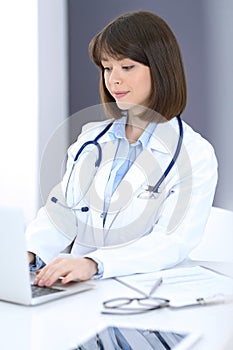 Doctor typing on laptop computer while sitting at the white table in hospital office. Physician at work. Medicine and