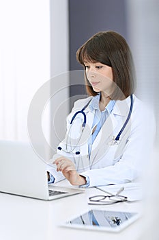 Doctor typing on laptop computer while sitting at the white table in hospital office. Physician at work. Medicine and