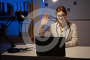 Doctor in uniform having video chat, consultation patient online sitting at table during night shift at hospital office.