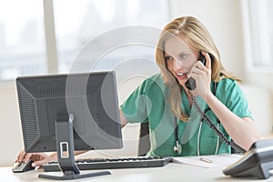 Doctor Using Computer While Conversing On Landline Phone At Desk