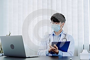 Doctor was sitting in the office working on a laptop computer while wearing a mask
