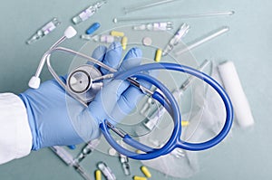 Doctor wearing blue gloves and holding stethoscope against pills, syringes, bandage on the blue medical table