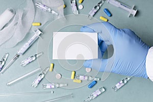 Doctor wearing blue medical gloves and holding empty business card against blue table. Pills,syringe, bandage as a background