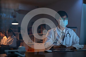 The doctor wearing mask is sitting at a desk at the night, Which is researching how to treat the patient`s condition.