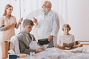 Doctor wearing medical apron and glasses talking with teenage patients sitting on hospital beds