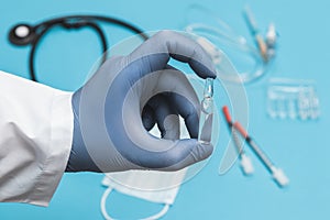 Doctor in a white coat holding glass ampoule. Hand in a blue sterilized surgical glove. Blurred background with medical tools