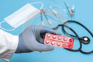 Doctor in a white coat holding tablets in red blister. Hand in a blue sterilized surgical glove. Blurred background with medical