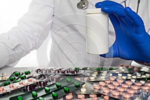 Doctor in white coat sits infront of pill boards scattered on table holding up blank prescription bottle.