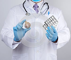 Doctor in a white medical coat and blue rubber gloves holds a fresh head of garlic and pills